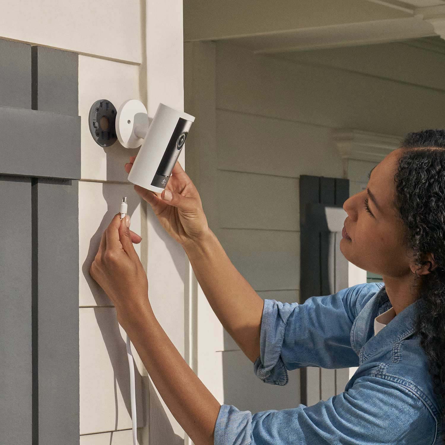 Outdoor Cam Pro (Featuring Retinal 4K) - Plug-In:Woman holding a white Ring Outdoor Cam Pro and power cord, installing the camera to a mount on an exterior wall.