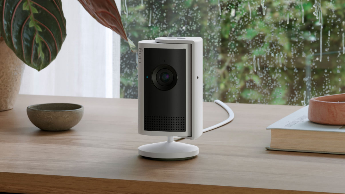 Compact white indoor camera on a wooden desk near a plant, bowl, and book, with a rainy window in the background.