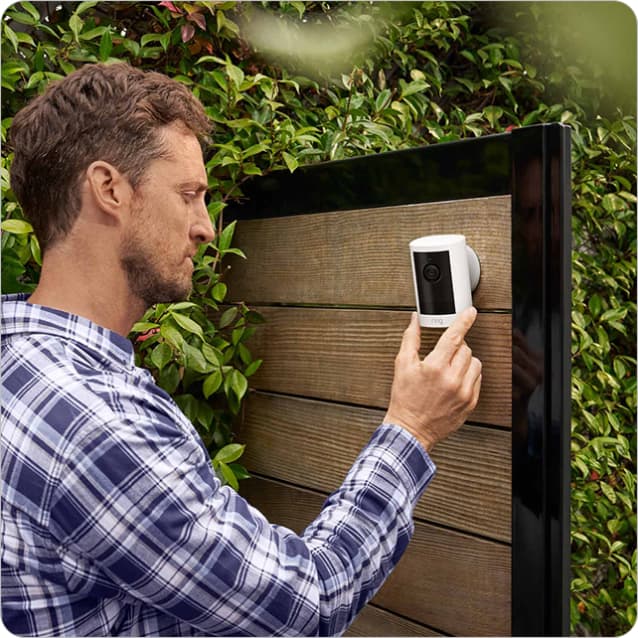 Person in plaid shirt mounts a white Ring outdoor camera on a wooden fence beside green foliage.