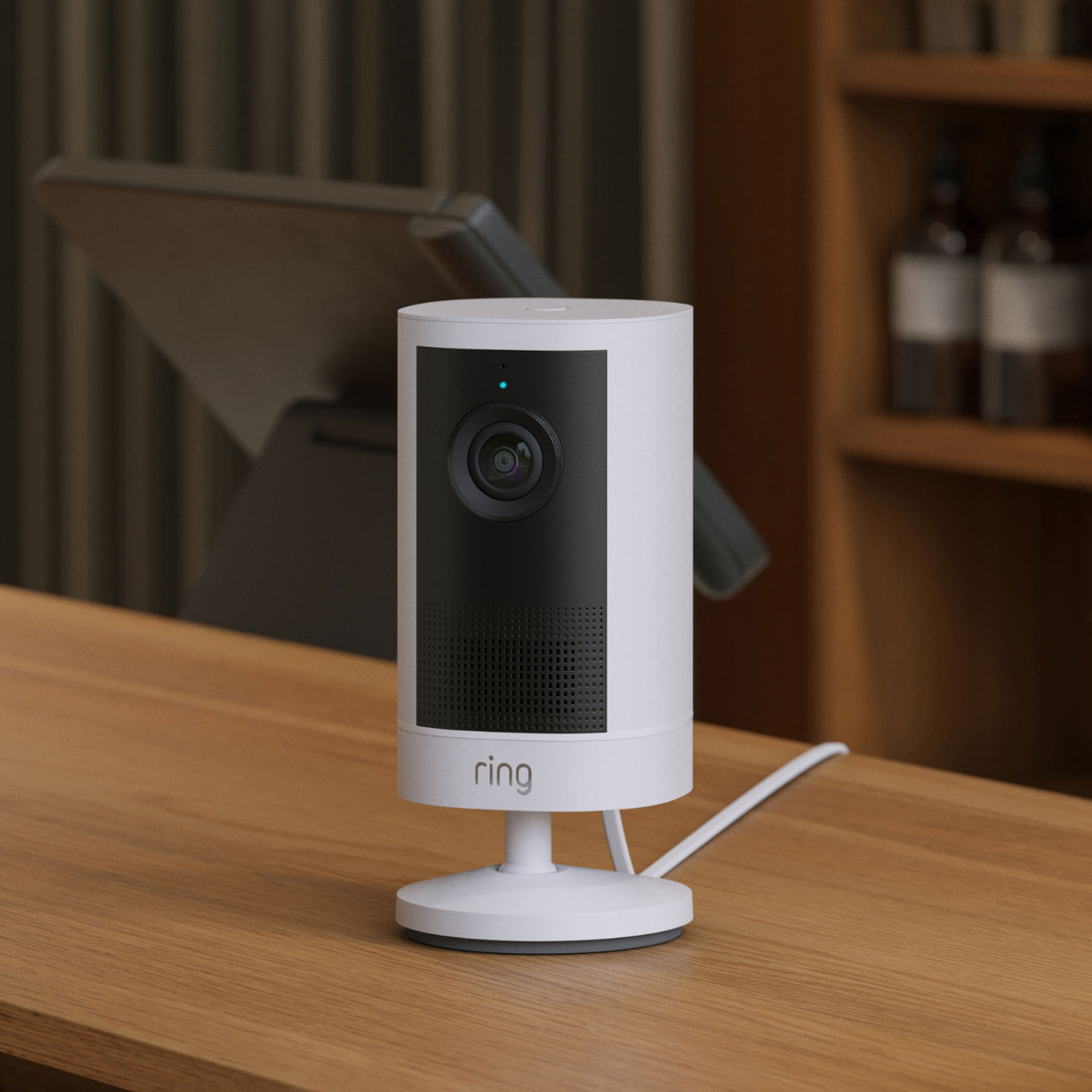 Outdoor Cam 4K Plus  - Plug-In:White Ring indoor camera on a wooden desk, cable plugged in, with a tablet stand and shelves blurred in the background.
