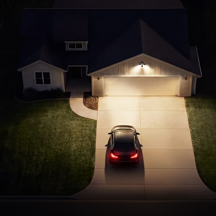 Nighttime view of a suburban house with a lit garage and driveway, a dark car parked with brake lights on near the entrance.