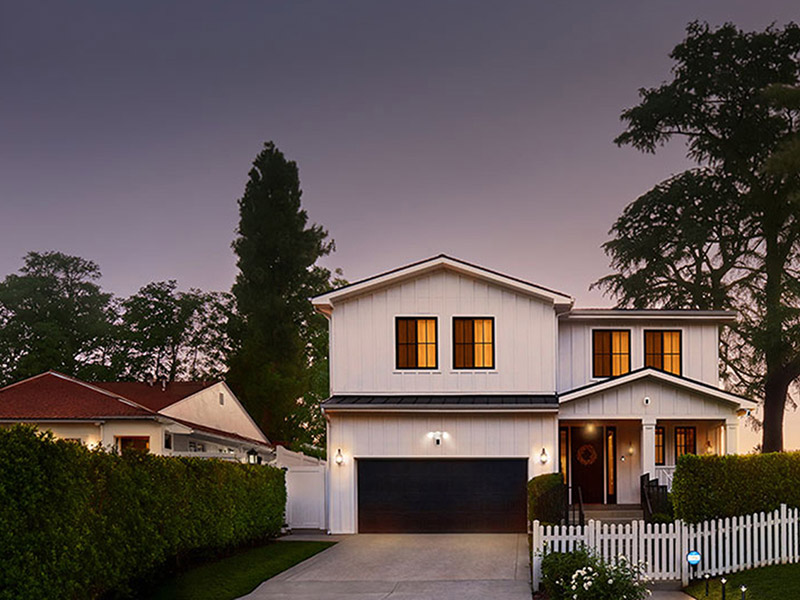 Two-story white house at dusk with warm-lit windows, black garage door, front porch, white picket fence, and trees in the background.