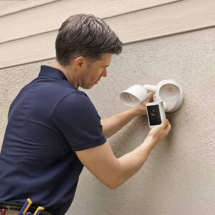 Person installing a white outdoor floodlight camera on a stucco wall, aligning the device beneath two lamp heads.