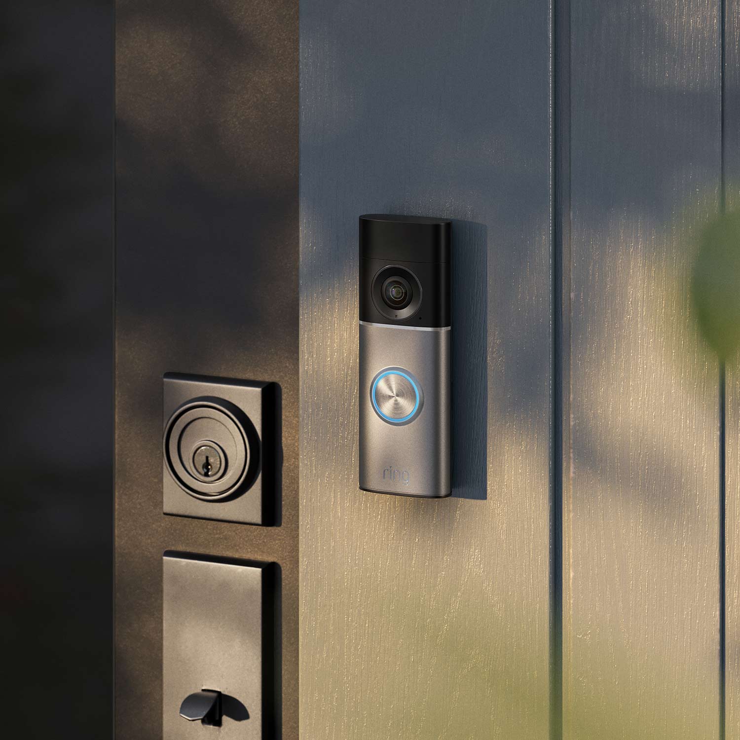 Smart video doorbell with glowing blue ring mounted on a gray front door beside a modern deadbolt and handle, lit by soft sunlight.