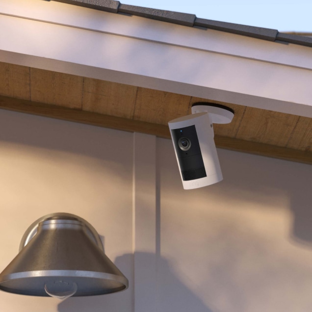 A white Ring Outdoor Cam Plus mounted under home's porch eaves, angled downward, next to a porch light.
