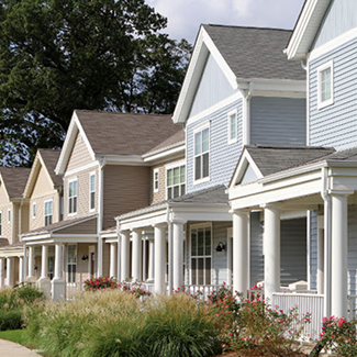 Row of suburban houses with front porches, pastel siding, manicured lawns, and flowers on a sunny day.