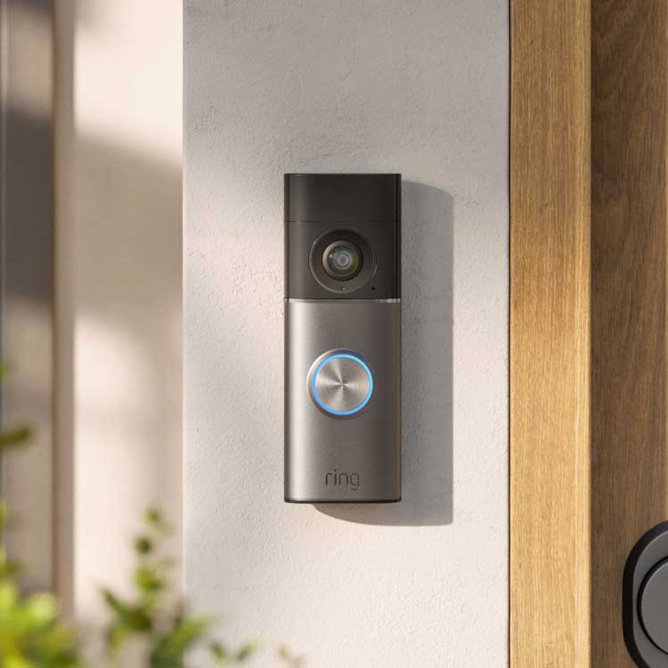 Smart doorbell with camera and glowing blue ring mounted on a light-colored wall beside a wooden doorframe in daylight.