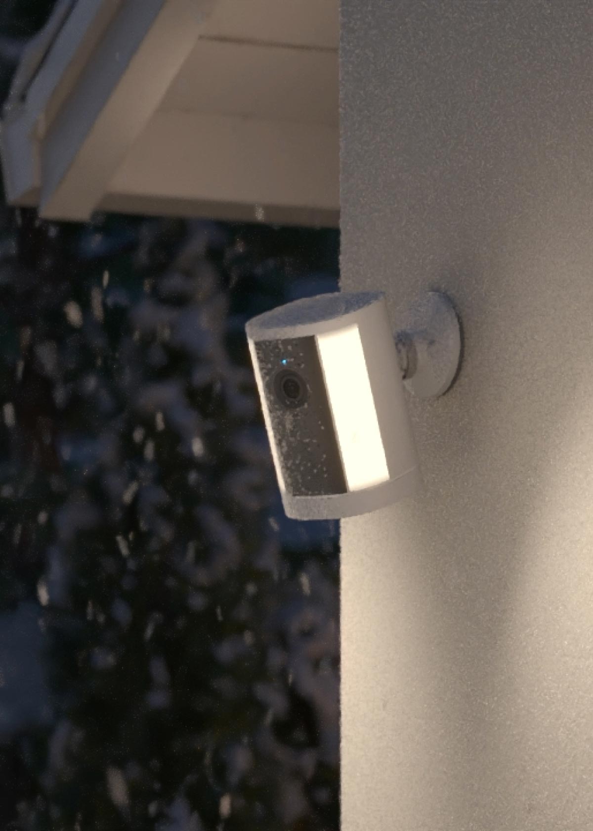 Outdoor security camera with a glowing light mounted on a frosty wall during snowfall near a house roofline at night.