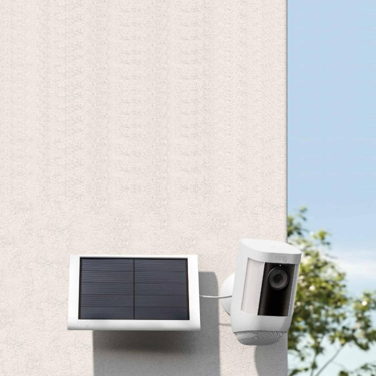 White Spotlight Cam Pro Battery camera connected to Solar Panel, mounted outside on textured natural colour wall. The day has blue sky and trees 