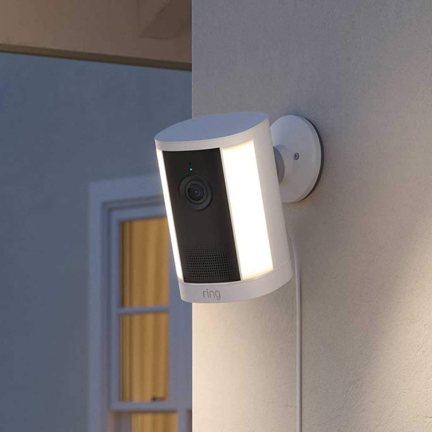 Spotlight Cam 4K Plus - Plug-In:White Ring outdoor camera with twin LED light strips mounted on a wall at dusk, cable running down, house window softly lit behind.