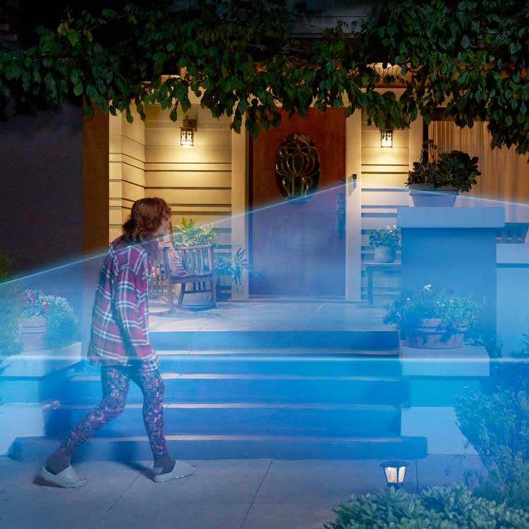 Night view of person in plaid approaching front porch, with decorative plants and exterior lights visible. A blue laser graphic shows the doorbell camera's detection zone.