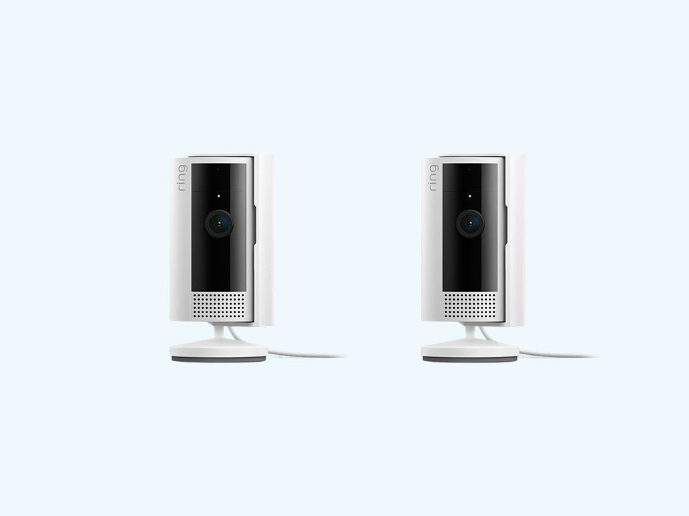 Two white Ring indoor security cameras on stands against a light blue background, showing black lens panels and speaker grilles.
