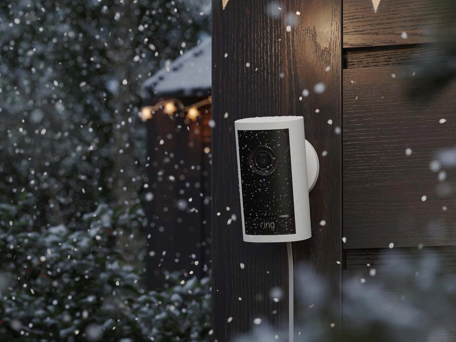 White outdoor cam pro mounted on an outdoor wooden wall, with snow falling in front.