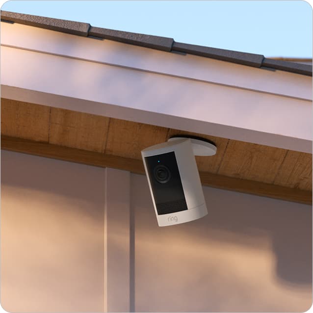 Outdoor Ring camera mounted under an eave near a metal porch light on a modern home at sunset.