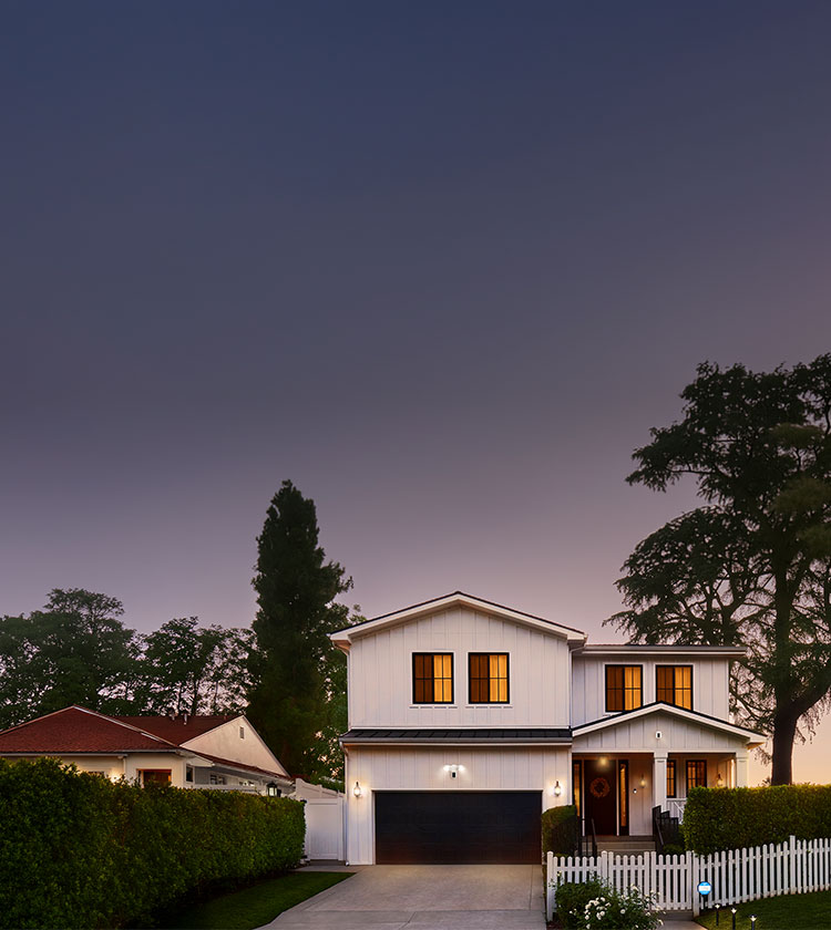 Two-story white house at dusk with warm interior lights, black garage door, white picket fence, hedges, and large trees in the yard.