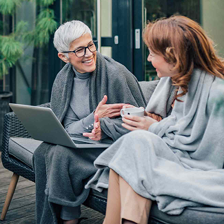 Two women on a patio sofa, wrapped in gray blankets, smiling and chatting over a laptop and a cup of coffee near greenery.