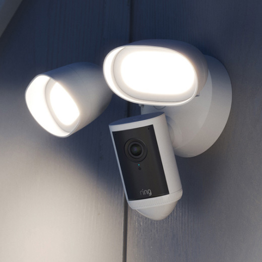 White Ring floodlight security camera mounted on an exterior wall at dusk, lens facing outward with a small blue status light.