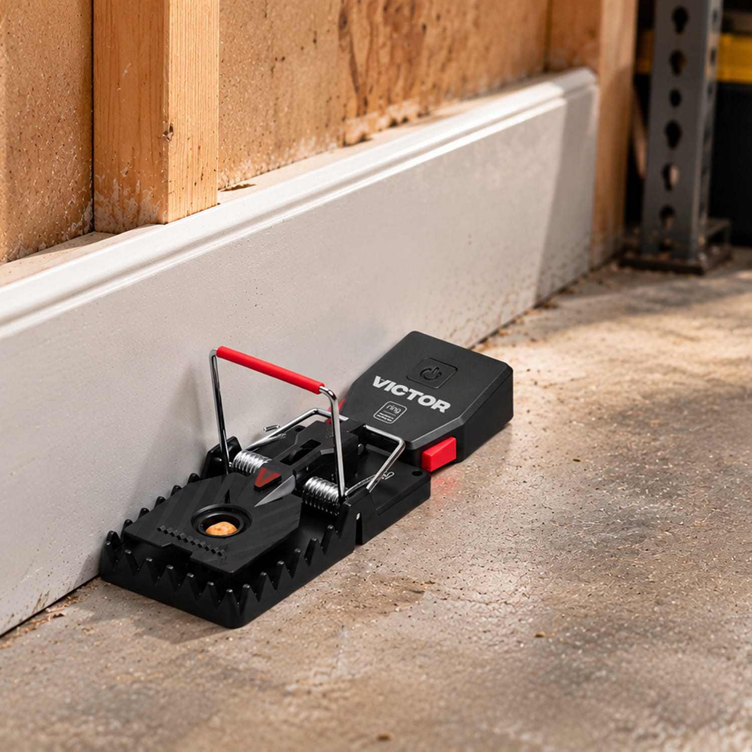 Victor® Smart Rat Trap, Indoor Snap Trap  - Black Victor rat trap with red accents set along a garage wall on a dusty concrete floor.