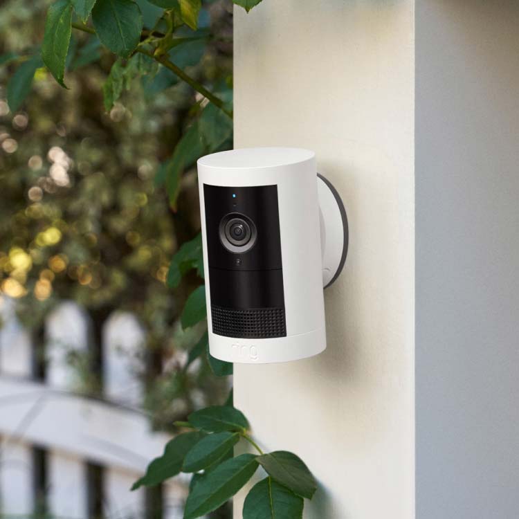 White outdoor security camera mounted on a wall near a garden and picket fence, with greenery in soft focus in the background.
