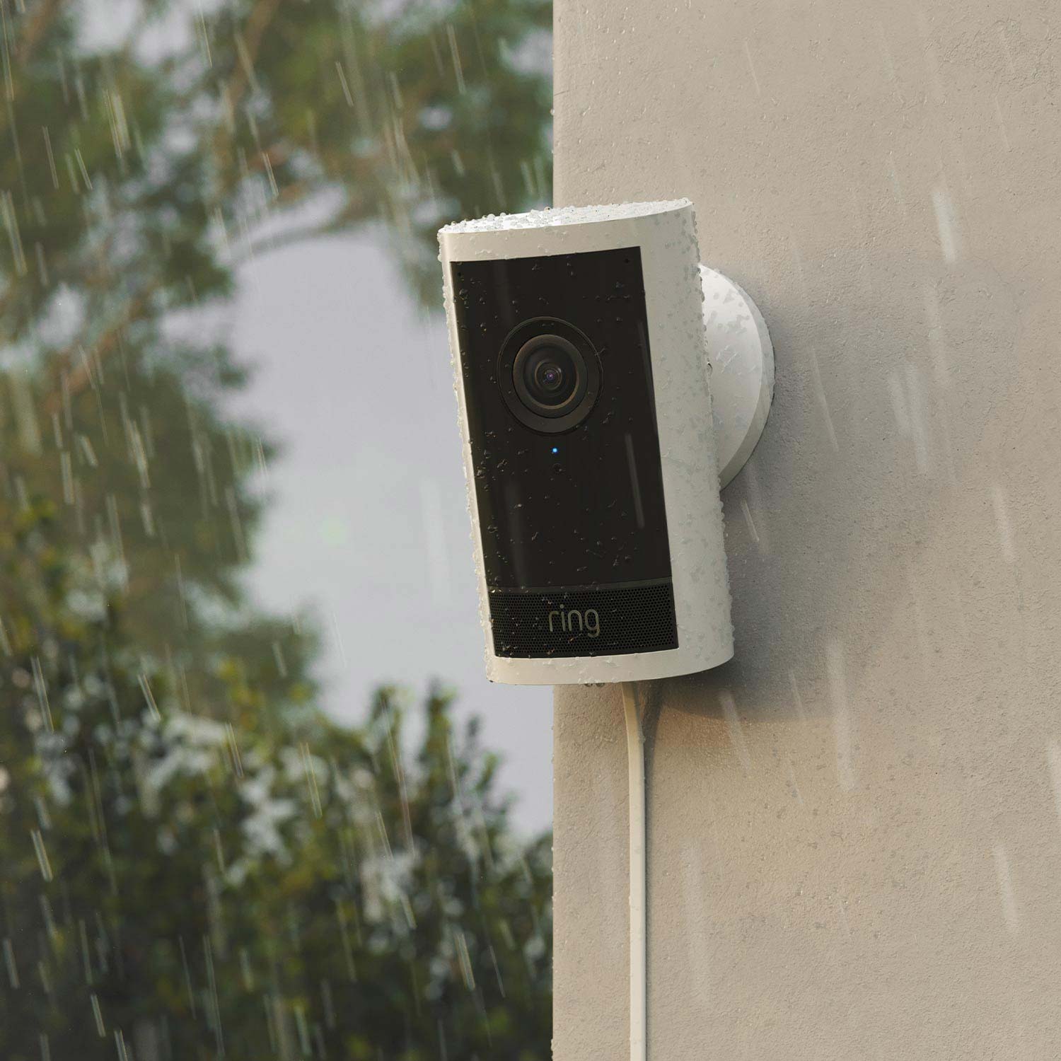 Outdoor Cam Pro (Featuring Retinal 4K) - Plug-In:White Ring Outdoor Cam Pro in the rain, mounted to a wall. Power cord stretches down the wall from the camera base.