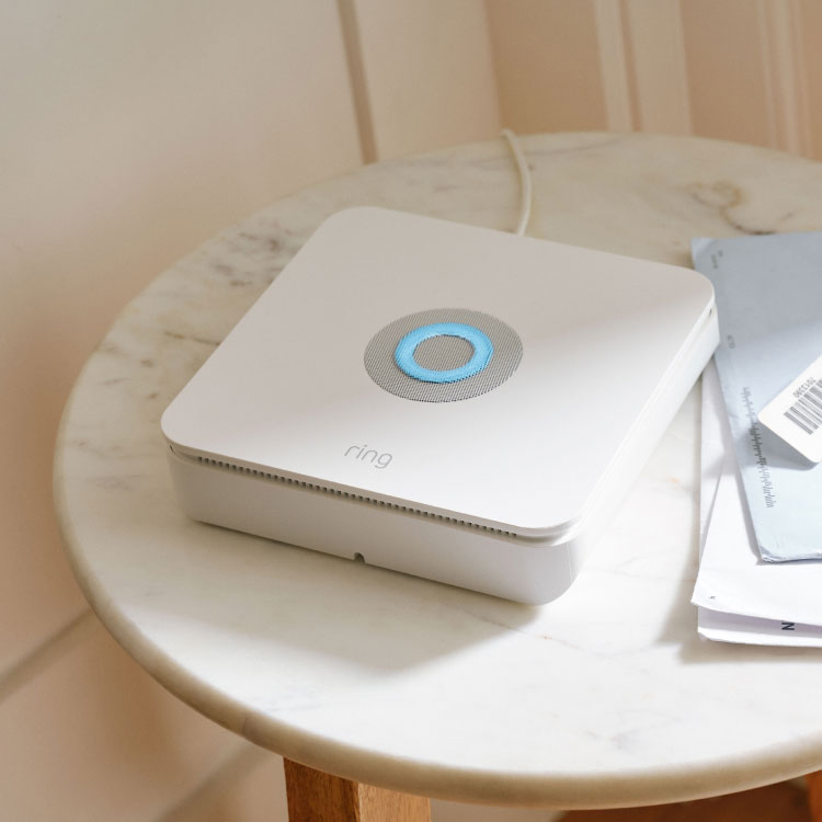 White Ring base station with blue-lit button on a marble table beside mail and a keyring, in a bright entryway.