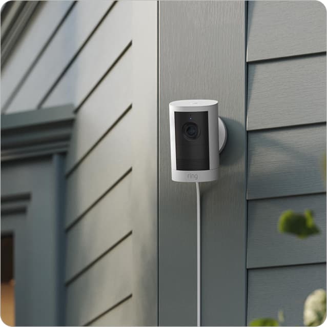 White Ring outdoor security camera mounted on gray house siding near a doorway, with power cable and soft daylight.