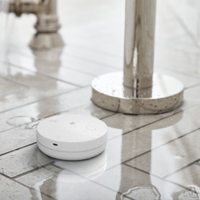 Small white smart water leak detector device on wet bathroom tile floor next to metal shower base.