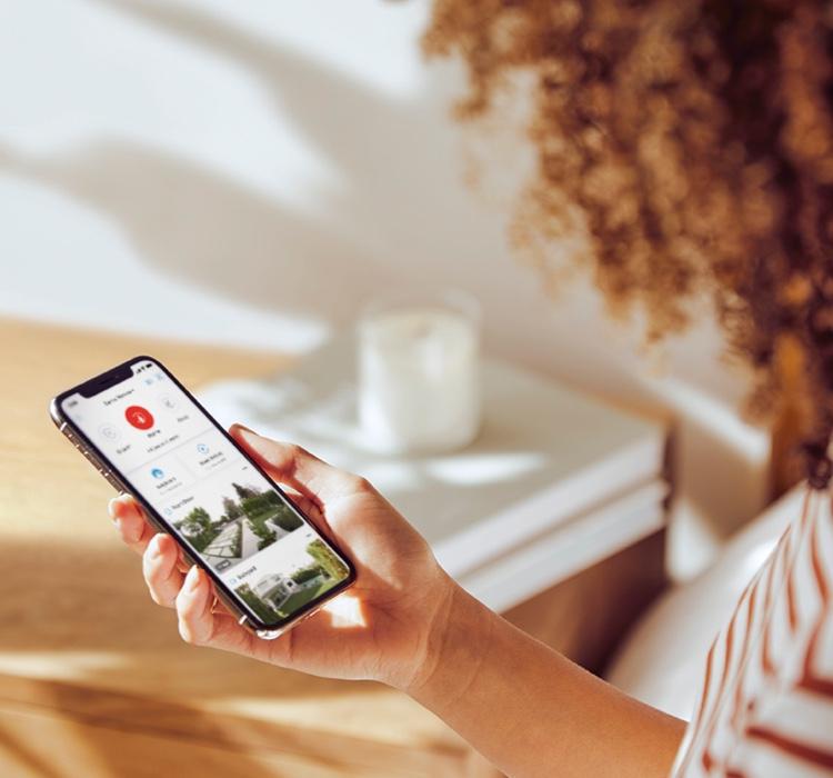 Woman with curly hair wearing a striped shirt holding a smartphone displaying the Ring App, monitoring her home security feed in a well-lit room.