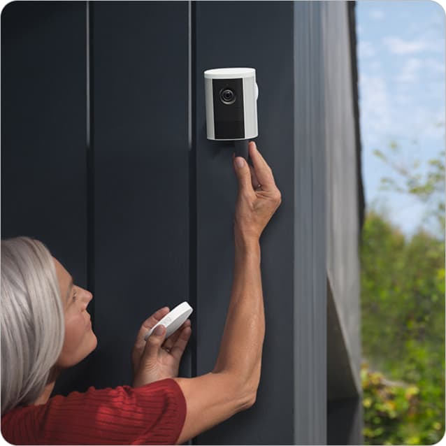 Person mounts a small Spotlight Cam (2nd Gen) on a wall, holding a tool, with greenery in the background.