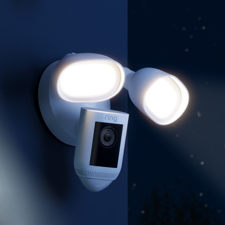 A black floodlight cam mounted over a garage shines brightly at dusk. 