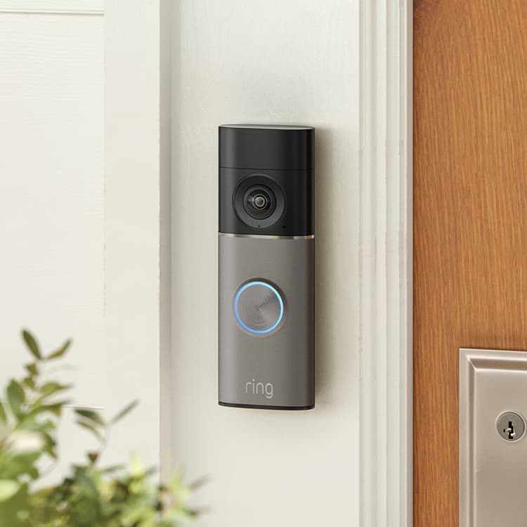 Video doorbell with camera and glowing blue ring mounted beside a wooden front door on a light-colored exterior wall.