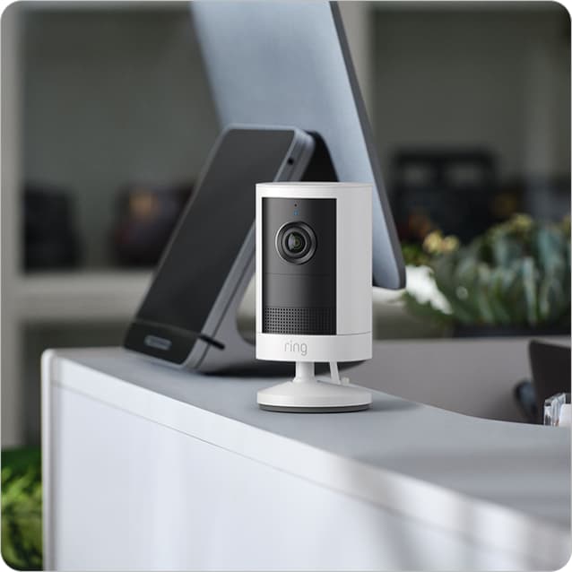 White Ring indoor camera on a desk near a tablet, with blurred shelves and plants in the background.