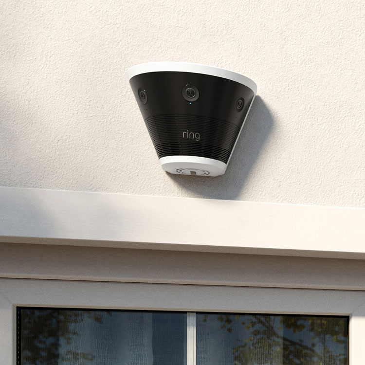 Ring outdoor camera mounted above a window on a light stucco wall, shown in daylight with reflections in the glass.