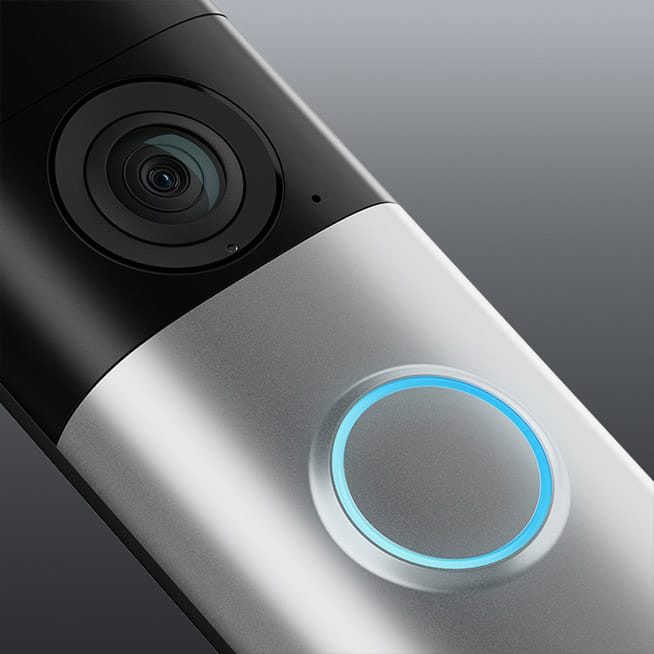 A close-up diagonal view of a Wired Doorbell Plus (2nd Gen) showing its sleek camera lens at the top and circular button with blue illuminated ring at the bottom against a grey background. Play button overlay is in the center of the image. 