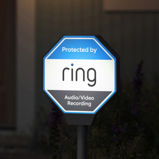 Illuminated blue-and-white yard sign reading “Protected by Ring” and “Audio/Video Recording” on a stake in a dimly lit front yard.