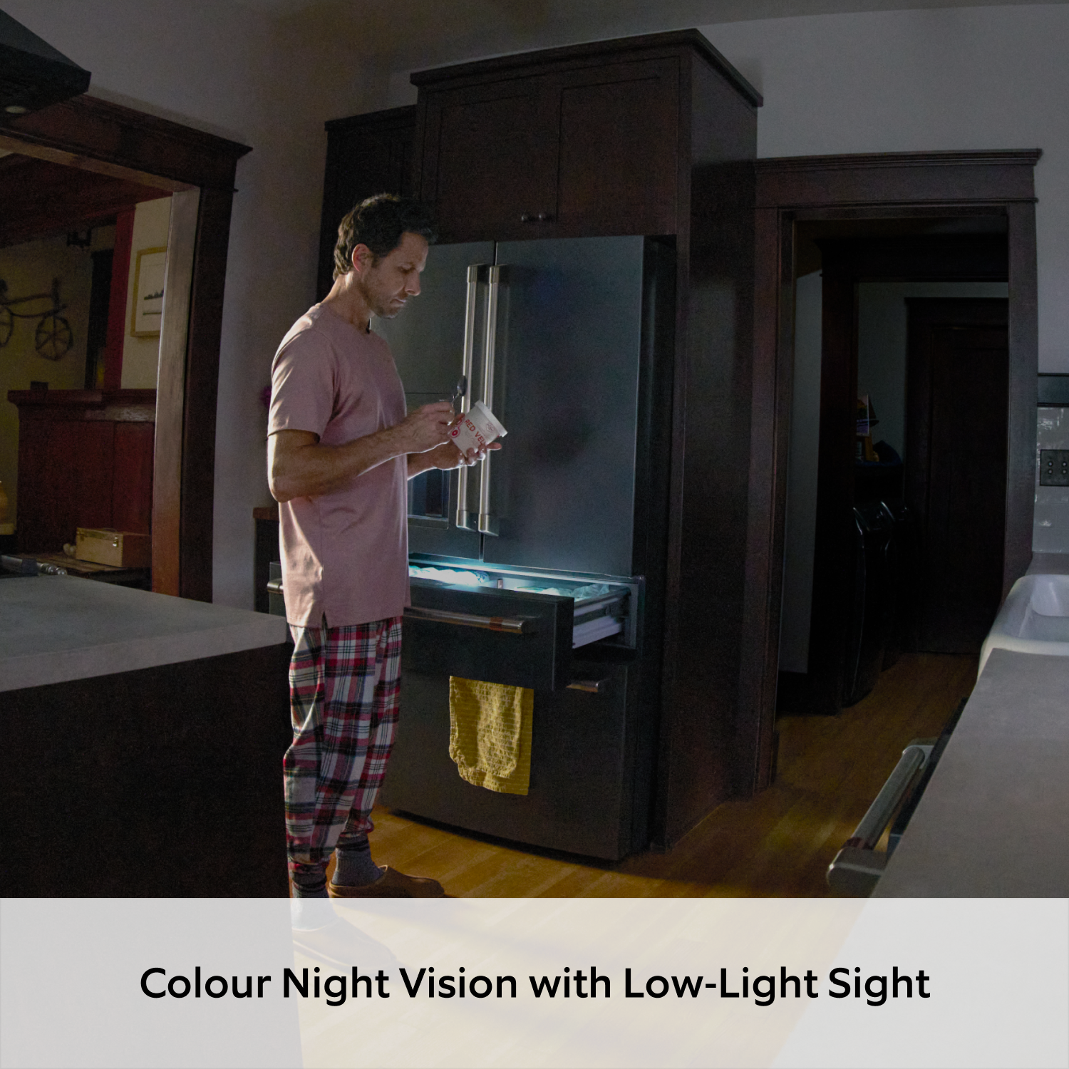 Indoor Camera Plus (Featuring Retinal 2K) - View of a home kitchen through an indoor security camera, showing a man in pyjamas standing by a refrigerator eating ice cream at night. The kitchen has dark cabinets, hardwood floors, and a ceiling fan visible in the frame.