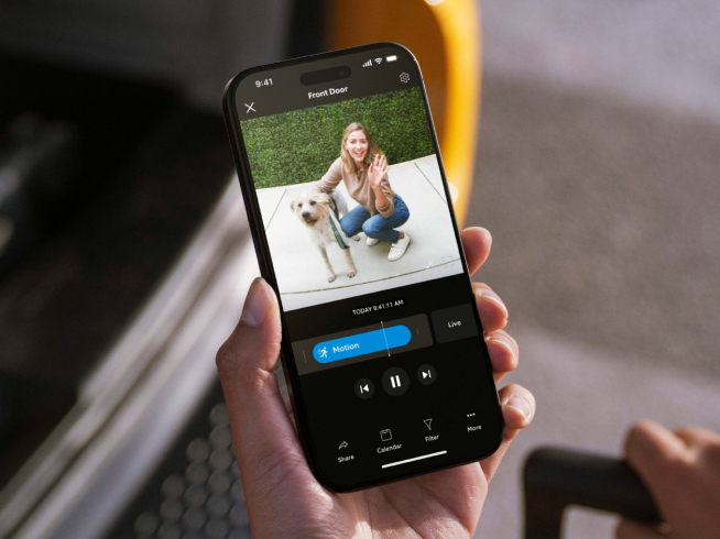 Hand holds smartphone displaying live Ring video feed of a smiling woman waving while crouching next to her dog.