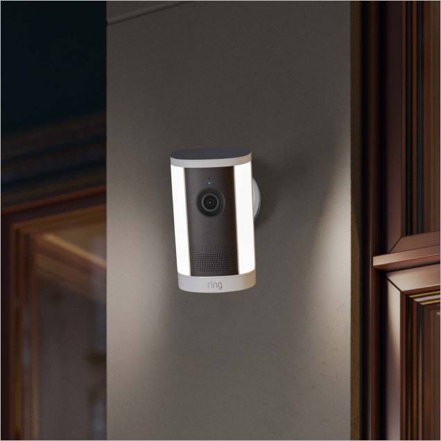 Wall-mounted Ring security camera with dual vertical LED lights glowing at dusk near a window frame.