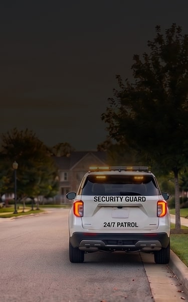 White SUV marked “Security Guard 24/7 Patrol” parked on a quiet suburban street at dusk, amber lights on, trees and homes nearby.