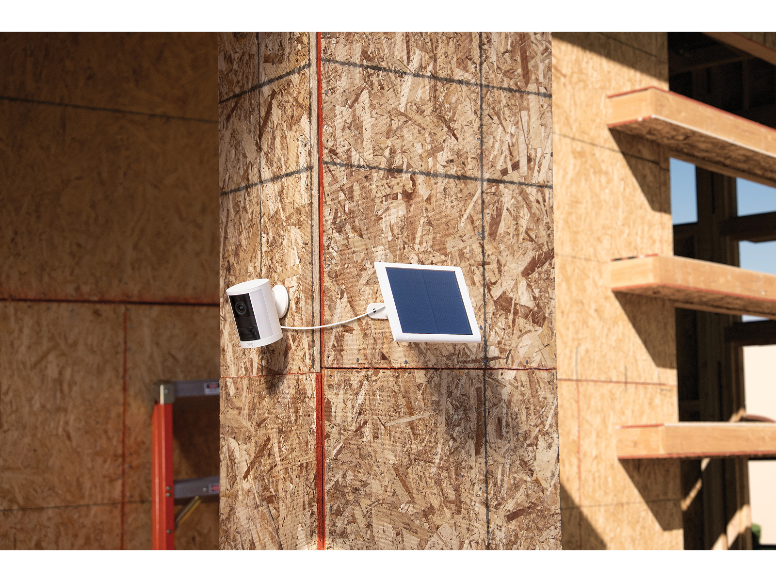 White outdoor camera and small solar panel mounted on an unfinished plywood wall at a construction site, connected by a short cable.