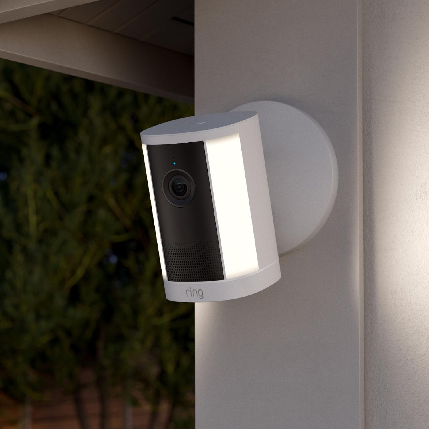 Spotlight Cam 4K Plus - Wired:White outdoor security camera with built-in light mounted on a wall under an eave at dusk, facing a yard with trees.