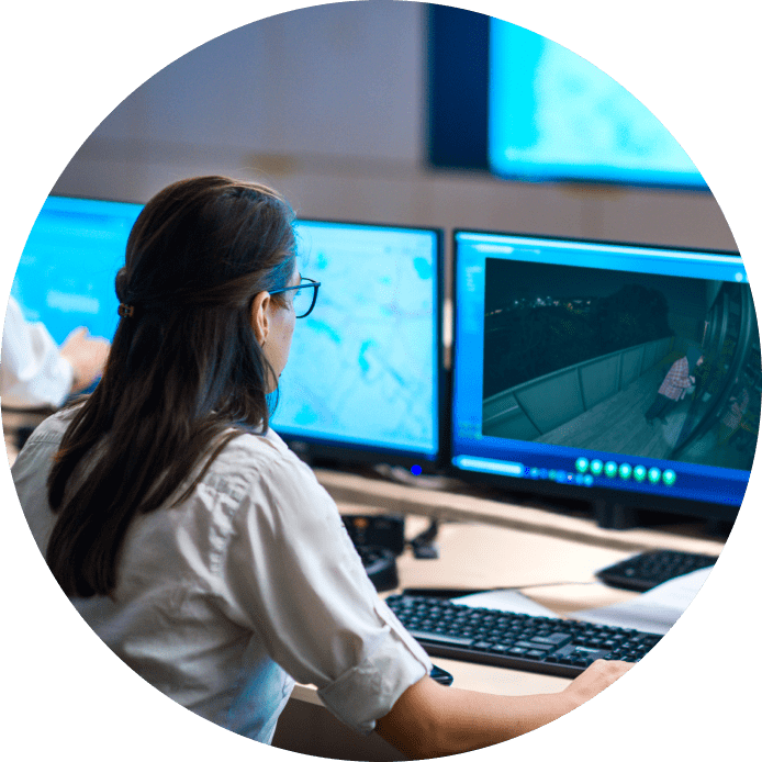 Person in white shirt working at a monitoring station with multiple blue-lit screens displaying data and surveillance footage.