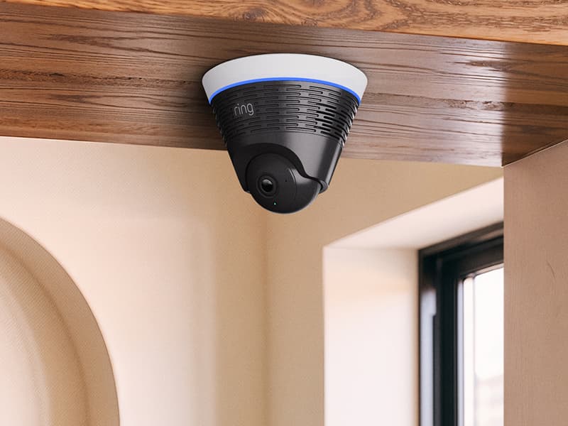 Black Ring indoor ceiling-mounted camera with blue light ring under a wooden beam in a bright room with a window nearby.