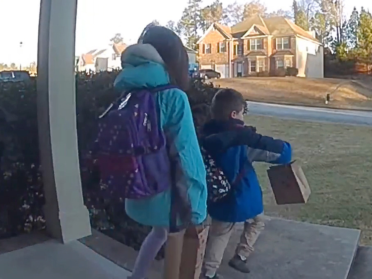 A young girl and boy wearing backpacks and holding brown paper bags walk off the front porch.