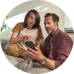 Teen and adult sitting on a couch, looking at a smartphone together in a bright living room with blankets and books in background.