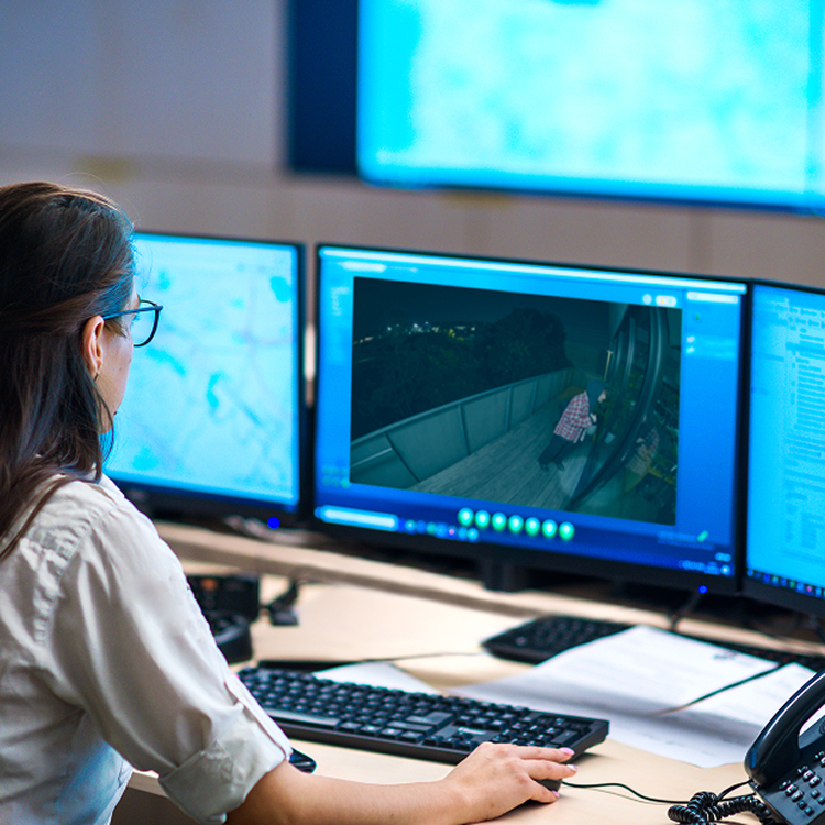 Operator at a desk reviews nighttime balcony footage on dual monitors in a control room, with keyboard, phone, and paperwork nearby.