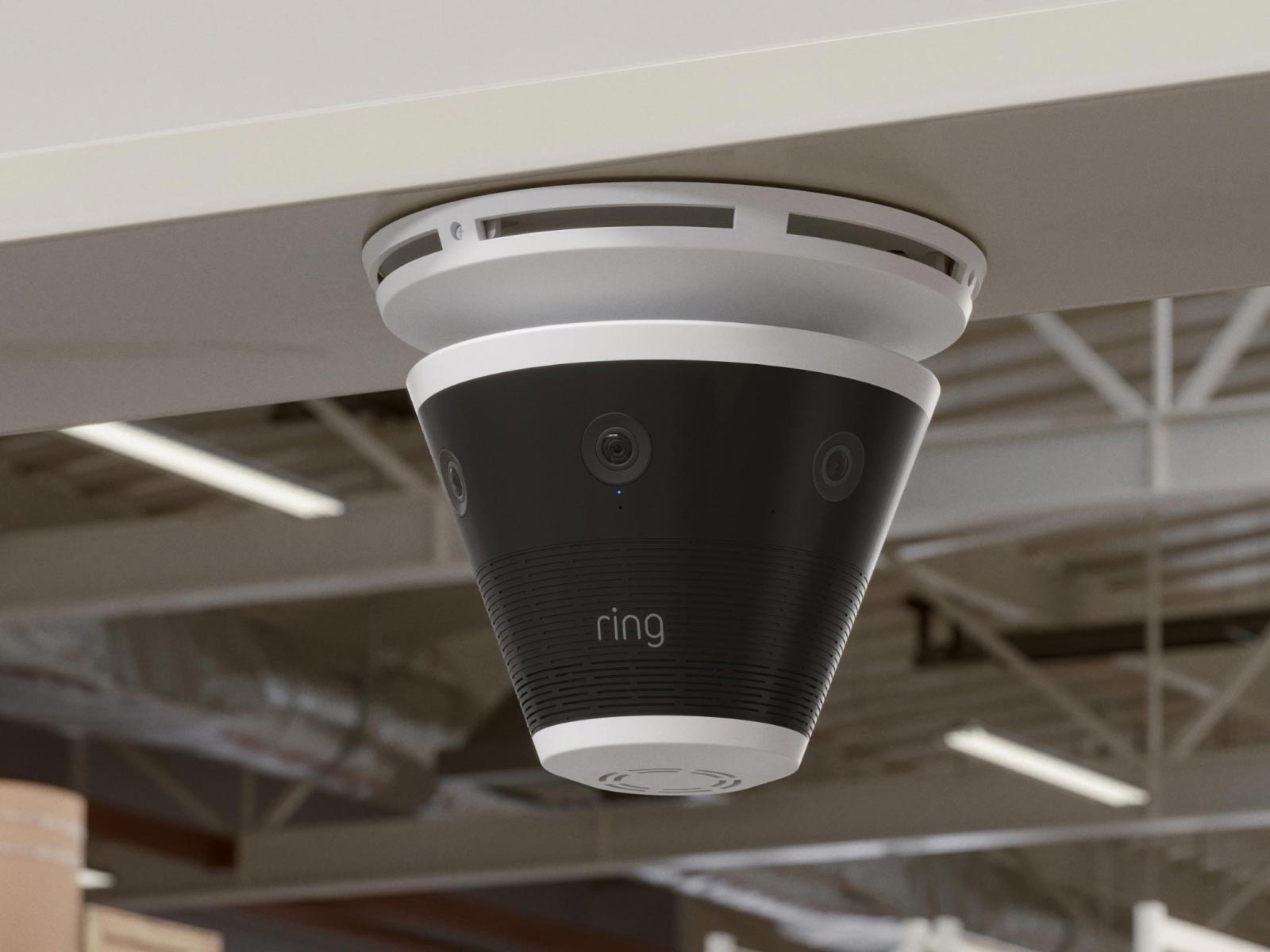 Ceiling-mounted Ring cone-shaped camera with multiple lenses in a warehouse-style interior, showing industrial beams and lights.