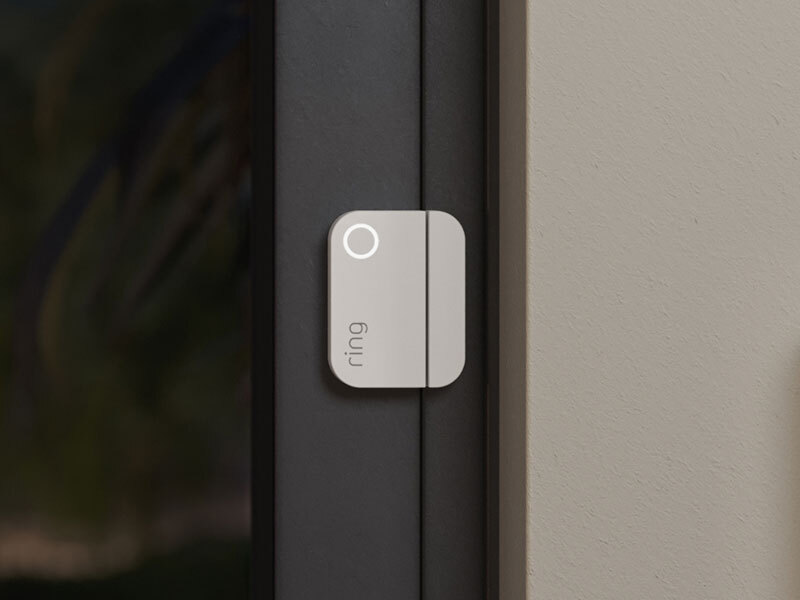Ring contact sensor with glowing LED mounted on a dark door frame next to a beige wall.