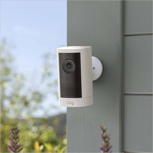 White Ring outdoor security camera mounted on a gray house wall, facing a garden with blurred plants and sky in the background.