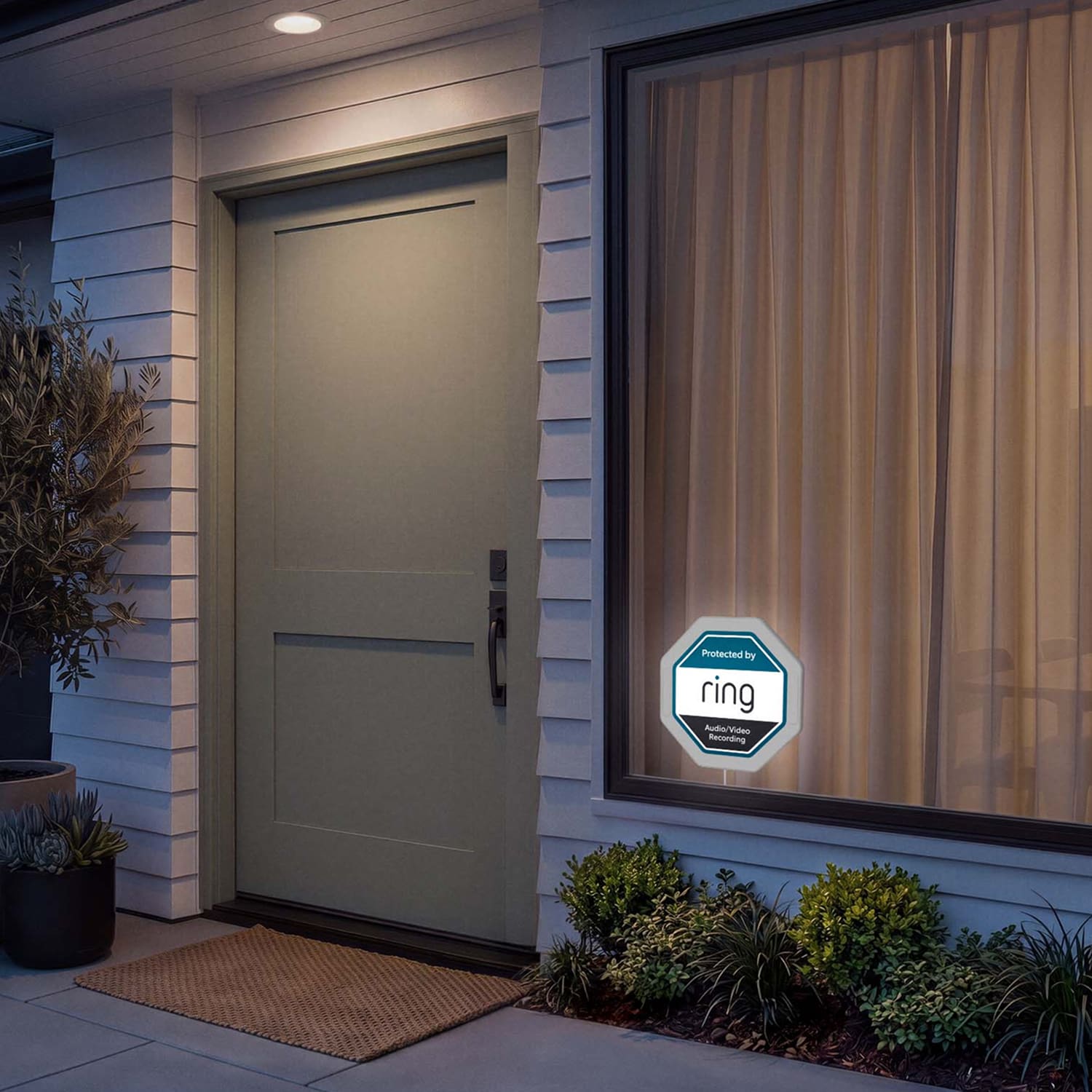 Ring Indoor Illuminated Sign - Evening view of a modern front entry with porch light on, beige door, window with curtains, Ring security sign, and small shrubs.
